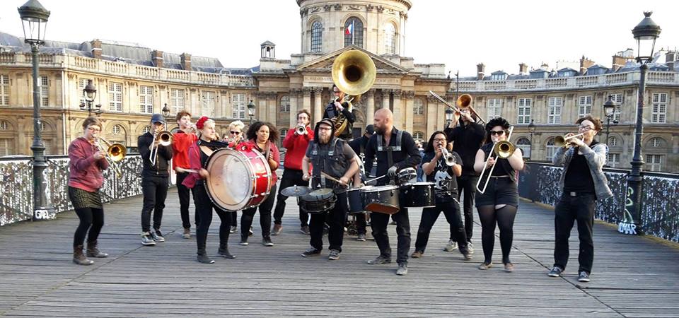 A fifteen member brass band with drums, horns, and trombones playing outdoors