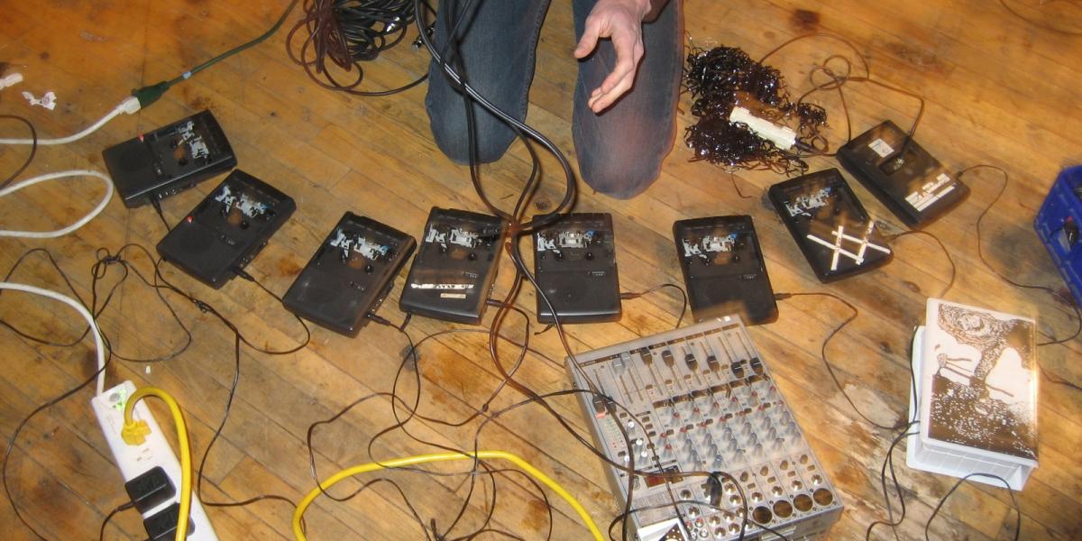 Performer kneels on a bare wood floor, surrounded by cables and cassette players