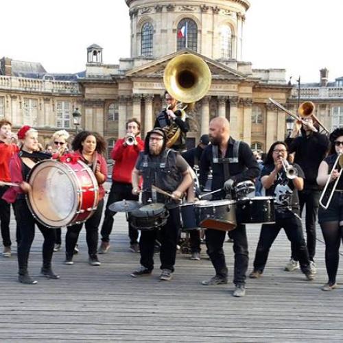 A fifteen member brass band with drums, horns, and trombones playing outdoors
